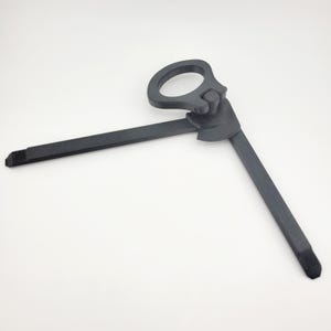 May include: A dark gray, hinged tool with a circular handle and two extending arms. The arms are rectangular and have flat, angled tips. The tool appears to be made of a solid, matte material.