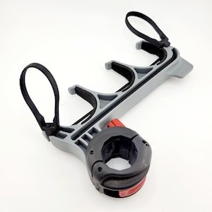May include: A gray and black bicycle accessory with a red accent. The accessory has a circular clamp, two curved supports, and two black straps. The item is designed to hold an object, such as a water bottle, on a bicycle.