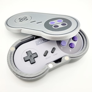 May include: A gray Super Nintendo controller case, open to reveal the controller inside. The case is shaped like a classic SNES controller, with a gray and purple color scheme. The text "SUPER NINTENDO ENTERTAINMENT SYSTEM" is visible.