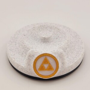 Op de afbeelding: Een wit en gespikkeld rond object met een zwarte basis en een kleine cilindrische top. Het heeft een goudkleurig embleem met een driehoekig ontwerp in een gouden cirkel. Waarschijnlijk een decoratief item of een game-accessoire.
