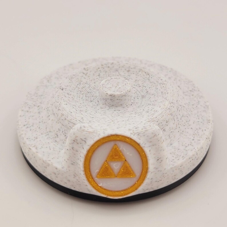 Op de afbeelding: Een wit en gespikkeld rond object met een zwarte basis en een kleine cilindrische top. Het heeft een goudkleurig embleem met een driehoekig ontwerp in een gouden cirkel. Waarschijnlijk een decoratief item of een game-accessoire.