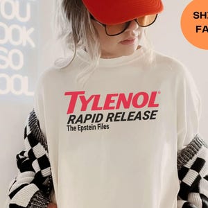 Może przedstawiać: Osoba nosząca białą koszulkę z nadrukiem "TYLENOL RAPID RELEASE" w kolorze czerwonym i czarnym. Osoba ta nosi również czerwoną czapkę typu trucker z napisem "MAD LOVE FOR YOU" w białym tekście. Czarno-biały sweter w kratę jest zarzucony na ramiona.