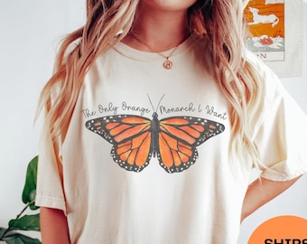 May include: Ivory-colored t-shirt featuring a large orange and black monarch butterfly graphic. The text above the butterfly reads "The Only Orange Monarch I Want." The shirt is paired with dark green shorts. An orange circle with the words "SHIPS FAST" is visible.