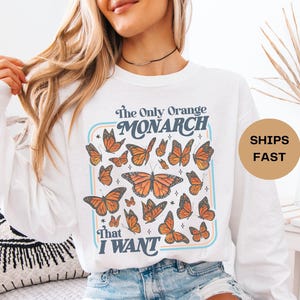 May include: White long-sleeve shirt with a graphic of orange monarch butterflies. The text on the shirt reads "The Only Orange Monarch That I Want" in a retro font. The shirt is paired with denim shorts.