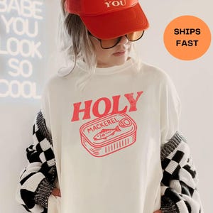 Puede incluir: Una persona lleva una gorra de camionero roja con el texto "MAD LOVE FOR YOU" y gafas de sol. Lleva una camiseta blanca con el texto "HOLY MACKEREL" y un gráfico rojo de una lata de sardinas. Un cárdigan a cuadros blanco y negro está sobre sus hombros.