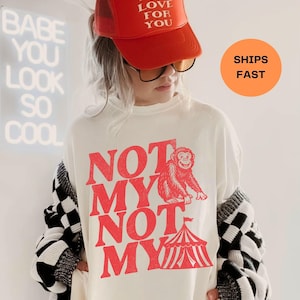 Puede incluir: Camiseta blanca con un gráfico rojo de un mono y el texto "NOT MY NOT MY". La persona lleva una gorra de camionero roja con el texto "MAD LOVE FOR YOU". Un cárdigan a cuadros blanco y negro está sobre los hombros.