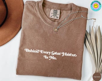 Może przedstawiać: Koszulka w kolorze espresso z napisem "Behind Every Great Haircut Is Me." Srebrny naszyjnik z wisiorkiem spoczywa na koszulce. Koszulka marki Comfort Colors, rozmiar S-CH.