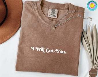 Może przedstawiać: Koszulka w kolorze espresso z białym napisem "I Will Cut You". Koszulka ma okrągły dekolt i krótkie rękawy. Srebrny naszyjnik z wisiorkiem spoczywa na koszulce.