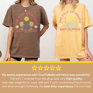 K&ouml;nnte beinhalten: Zwei T-Shirts mit einem Vintage-Grafikdesign mit einer Palme und dem Text "The Gulf of Mexico" und "Golfo de Mexico Est. 1550". Die Shirts sind in zwei Farben erh&auml;ltlich: Espresso-Braun und Senfgelb.