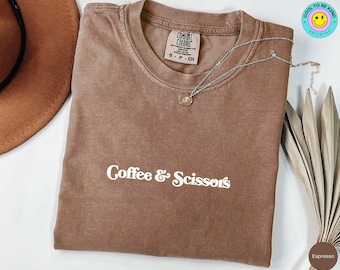 Może przedstawiać: Koszulka w kolorze espresso z napisem "Coffee & Scissors" w białym skrypcie. Koszulka ma okrągły dekolt i krótkie rękawy. Srebrny naszyjnik z małą zawieszką spoczywa na koszulce. Brązowy kapelusz i suszone liście znajdują się w tle.