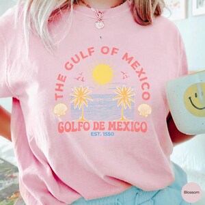 K&ouml;nnte beinhalten: Rosafarbenes T-Shirt mit einem grafischen Design aus Palmen, einer Sonne und dem Text "The Gulf of Mexico, Golfo de Mexico, Est. 1550".