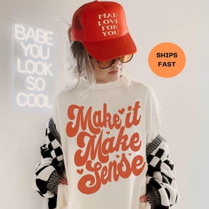 以下が含まれることがあります： オレンジ色のスクリプトで「Make it Make Sense」と書かれた白いTシャツ。赤いトラッカーハットには「MAD LOVE FOR YOU」とプリントされています。白と黒のチェック柄のカーディガンが肩にかけられています。