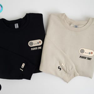 May include: Two long-sleeved sweatshirts, one black and one sand-colored, are displayed. Each features a retro video game controller graphic with the text "Player One" and "Player Two." The black sweatshirt has a controller graphic on the sleeve.