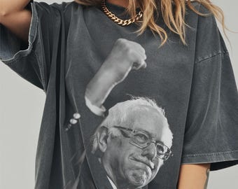 May include: Dark gray t-shirt featuring a black and white image of a man wearing glasses and a suit, with a raised fist above his head. The shirt has a vintage, worn look.