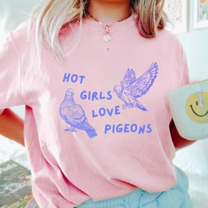 Może przedstawiać: R&oacute;żowa koszulka z niebieskim nadrukiem dw&oacute;ch gołębi i tekstem "Hot Girls Love Pigeons".