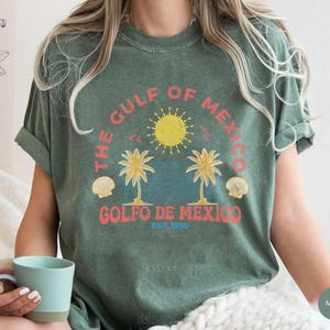 K&ouml;nnte beinhalten: Ein gr&uuml;nes T-Shirt mit einem Vintage-Grafikdruck von Palmen, einer Sonne und Muscheln. Der Text auf dem Shirt lautet "The Gulf of Mexico, Golfo de Mexico, Est. 1950".