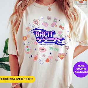 May include: Cream-coloured t-shirt featuring a vibrant graphic design. The design incorporates the text "BACH TO THE 2K" and "EMILY'S BACH PARTY", alongside various Y2K-themed icons. The shirt also has a yellow banner that says "PERSONALIZED TEXT!" and a purple circle that says "MORE COLORS AVAILABLE".