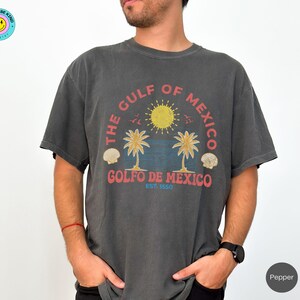 K&ouml;nnte beinhalten: Ein dunkelgraues T-Shirt mit einem grafischen Aufdruck von Palmen und einer Sonne. Der Text "The Gulf of Mexico" und "Golfo de Mexico Est. 1550" ist auf dem T-Shirt aufgedruckt.
