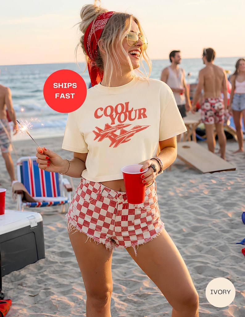 Pu&ograve; includere: T-shirt color avorio con la scritta "Cool Your" e grafica di un jet rosso. La modella indossa pantaloncini a scacchi rossi e bianchi e una bandana rossa. Lo sfondo mostra una scena sulla spiaggia con persone e un bicchiere rosso.