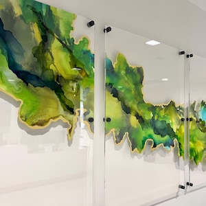 May include: Abstract wall art featuring three clear acrylic panels with vibrant green, yellow, and blue ink designs. The edges are outlined in gold, and the panels are mounted with black hardware, creating a modern aesthetic.