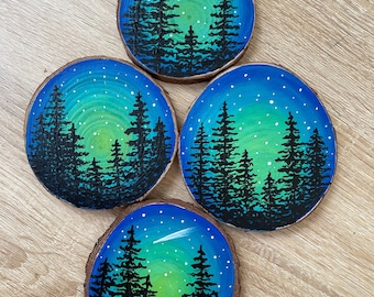 May include: Four round wood slices painted with a night sky scene. Each slice features a dark green and blue gradient sky with white stars and a shooting star. Silhouettes of pine trees are in the foreground.