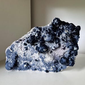 May include: A large, irregularly shaped mineral specimen with a textured surface. The stone features a mix of deep blue, light blue, and white colors. Dark blue, spherical formations are clustered across the surface, creating a unique, natural aesthetic.