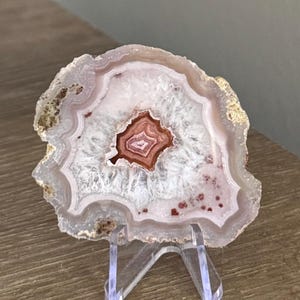 May include: A polished agate slice featuring concentric bands of pale pink, white, and translucent crystalline layers. The center displays a deep reddish-brown core with distinct banding, while the outer edges show a rough, natural mineral texture.