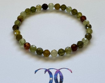 May include: A beaded bracelet featuring faceted gemstones in shades of green, brown, and clear. The bracelet is circular and rests on a white surface. A logo with two interlocking circles is at the bottom.