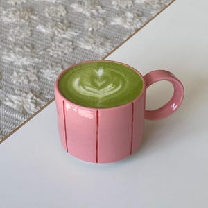 Könnte beinhalten: Ein rosa Keramikbecher mit einem abgerundeten Henkel und vertikalen roten Linien, gefüllt mit einem leuchtend grünen Matcha-Latte, der ein weißes, herzförmiges Schaummuster auf der Oberfläche aufweist. Der Becher hat eine glatte, glänzende Oberfläche.