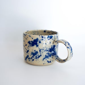 Könnte beinhalten: Eine Keramiktasse mit cremefarbenem Grund und einem gesprenkelten blauen Farbmuster. Die Tasse hat einen abgerundeten Henkel und eine zylindrische Form. Die Tasse eignet sich für Kaffee, Tee oder andere Getränke.