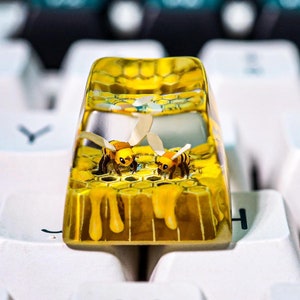 May include: A custom keyboard keycap with a yellow honeycomb design and two cartoon bees. The keycap is set against a background of white keyboard keys. A unique accessory for mechanical keyboards.