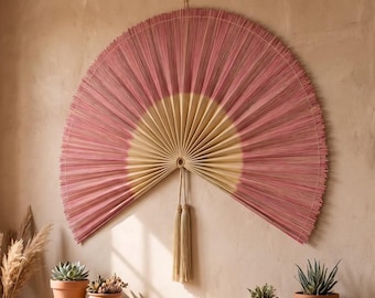 May include: A large, semi-circular wall fan made of thin, radiating sticks. The inner portion is a light beige, while the outer edge is a dusty rose pink. Two long, matching beige tassels hang from the center point of the fan's base.