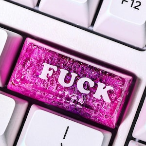 May include: Close-up of a white computer keyboard with a custom pink key that reads "FUCK" in white capital letters. The key is surrounded by other white keys with function keys and symbols.