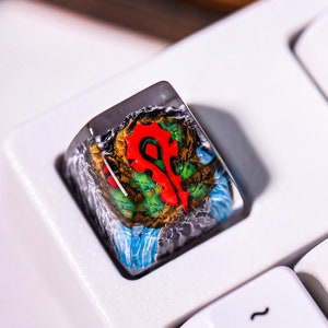 May include: A custom keycap featuring the Horde symbol from World of Warcraft. The keycap is clear with a colorful design, including red, green, and blue elements. It is set on a white keyboard, with other keys visible.