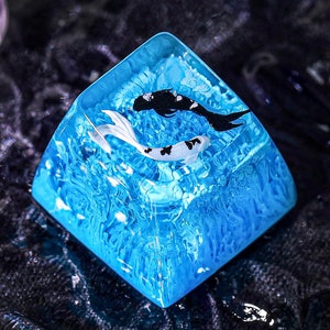 Op de afbeelding: Een vierkant, blauw kunstwerk van hars met twee koi-vissen. Eén koi is zwart-wit, de andere is zwart. De hars heeft een waterachtige textuur. Het kunstwerk staat op een donkere, getextureerde ondergrond.