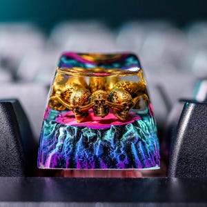 May include: A custom keycap with a skull design. The keycap is made of clear resin, with a gold skull design inside. The base of the keycap has a colorful, textured design with shades of blue, purple, and pink. The keycap is on a black keyboard.