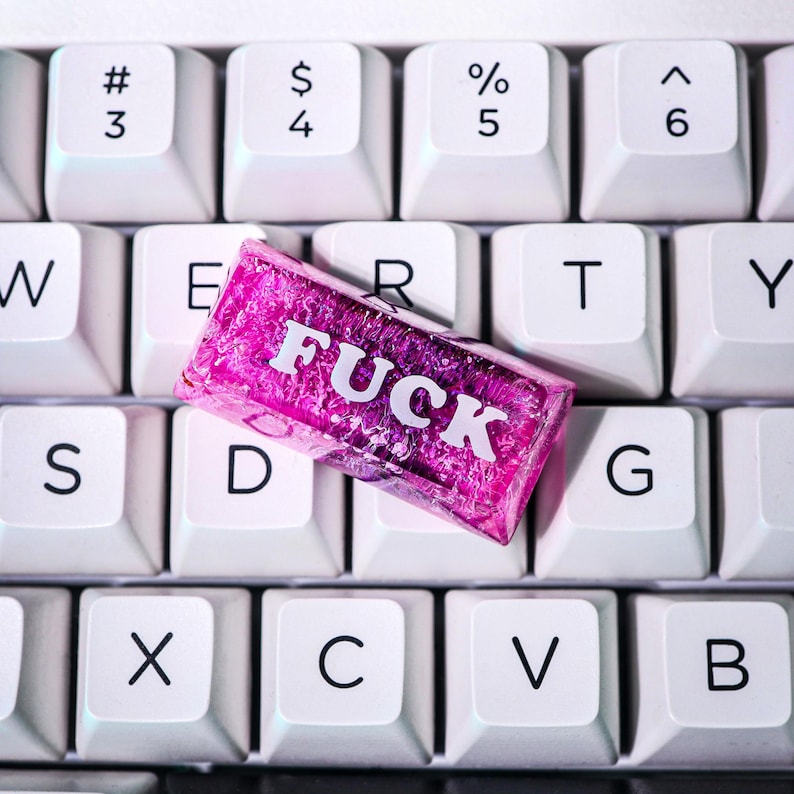 May include: A close-up of a white keyboard with a pink candy bar that reads "FUCK" in white capital letters. The candy bar is resting on the keys. The keyboard keys are arranged in a standard QWERTY layout.