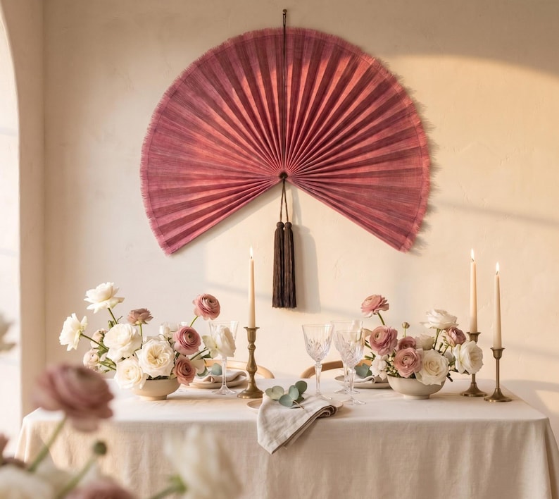 May include: A large, semi-circular, pleated wall fan in a deep dusty rose color, featuring a dark brown tassel hanging from the center. The fan is mounted on a wall above a dining table set with white and pink flowers, candles, and glassware.