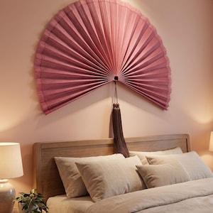 May include: A large, semi-circular wall fan in a dusty rose color with fine, vertical pleats radiating from a central point. A long, dark brown decorative tassel hangs from the center of the fan, which is mounted on a solid wall surface.