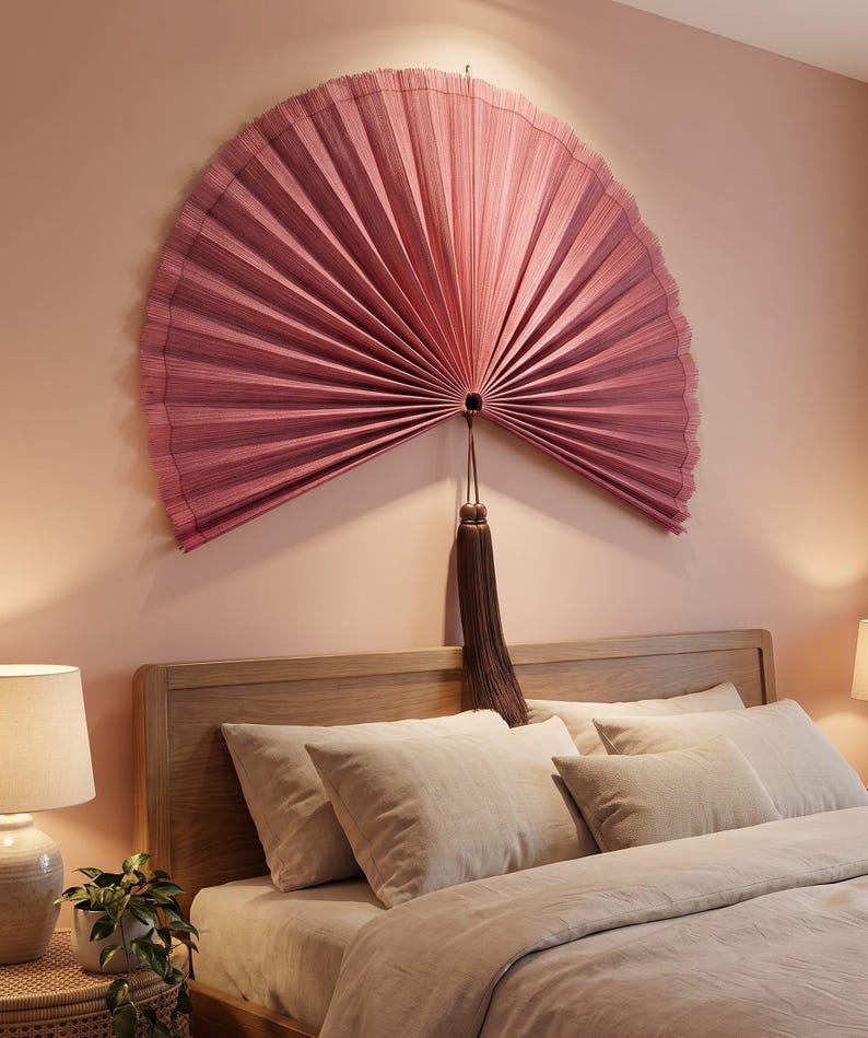May include: A large, semi-circular wall fan in a dusty rose color with fine, vertical pleats radiating from a central point. A long, dark brown decorative tassel hangs from the center of the fan, which is mounted on a solid wall surface.