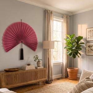 May include: A large, semi-circular, pleated wall fan in a deep rose-pink color, featuring a textured fabric surface and a decorative black tassel hanging from the center point, mounted on a light gray wall above a wooden slatted media console.