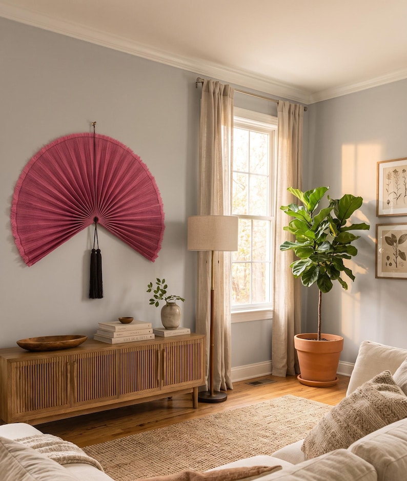 May include: A large, semi-circular, pleated wall fan in a deep rose-pink color, featuring a textured fabric surface and a decorative black tassel hanging from the center point, mounted on a light gray wall above a wooden slatted media console.