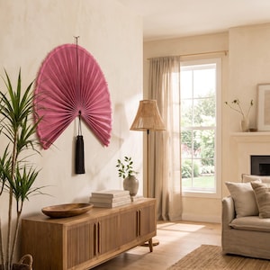 May include: A large, semi-circular wall fan in a vibrant magenta pink hue, featuring a pleated texture and a fringed edge. The fan is suspended from a central point and is accented by a long, dark black tassel hanging from the bottom center.
