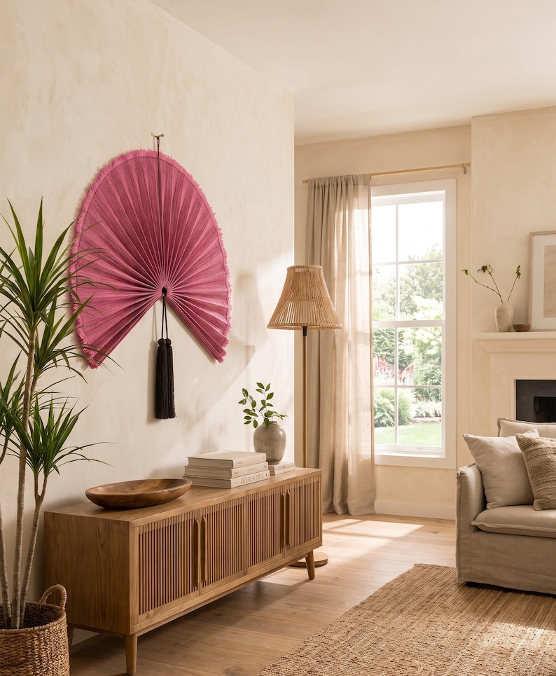 May include: A large, semi-circular wall fan in a vibrant magenta pink hue, featuring a pleated texture and a fringed edge. The fan is suspended from a central point and is accented by a long, dark black tassel hanging from the bottom center.