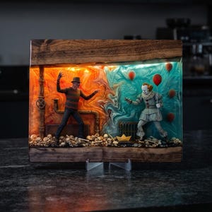 Op de afbeelding: Een rechthoekig houten diorama met twee horrorfiguren in hars. Links staat een figuur in een gestreepte trui in oranje licht. Rechts houdt een clownfiguur een rode ballon vast in blauw licht. Schedels liggen onderaan het diorama.