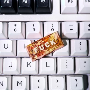 May include: A rectangular, amber-colored resin art piece with gold flakes and the word "FUCK" in white capital letters, resting on a white keyboard. The keyboard has black and white keys.