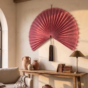May include: A large, semi-circular wall fan in a deep dusty rose color with a pleated texture and frayed edges. The fan is suspended by a thin cord and features two long, dark brown decorative tassels hanging from the center point of the fan.
