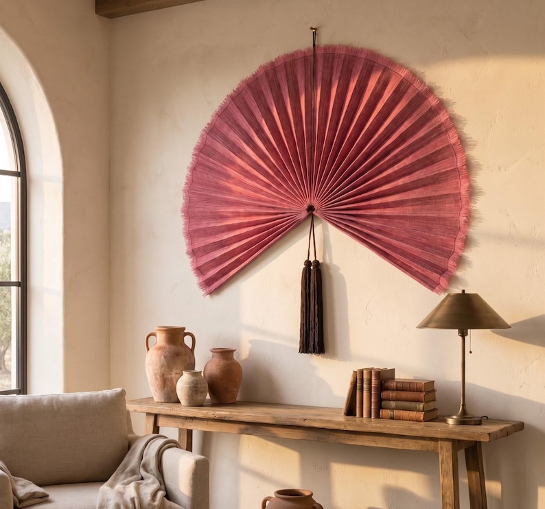 May include: A large, semi-circular wall fan in a deep dusty rose color with a pleated texture and frayed edges. The fan is suspended by a thin cord and features two long, dark brown decorative tassels hanging from the center point of the fan.