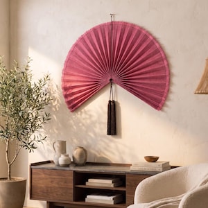 May include: A large, semi-circular wall fan in a deep magenta hue with a pleated texture and frayed edges. The fan is suspended by a thin cord and features a long, dark brown decorative tassel hanging from the center point of the base.