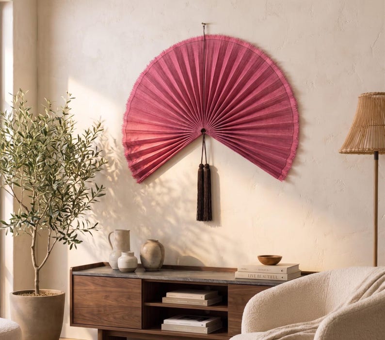 May include: A large, semi-circular wall fan in a deep magenta hue with a pleated texture and frayed edges. The fan is suspended by a thin cord and features a long, dark brown decorative tassel hanging from the center point of the base.